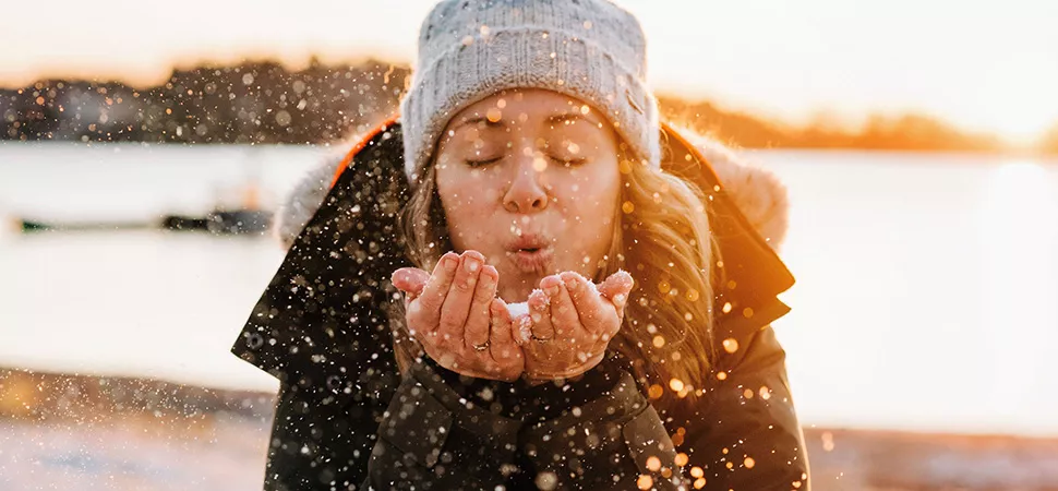 Eine Frau in Winterjacke & Mütze pustet Schnee in die Kamera 