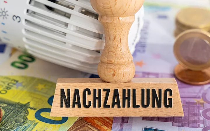 Geldscheine übereinander, darauf ein Heizungsthermostat und ein Stempel mit der Aufschrift "Nachzahlung"