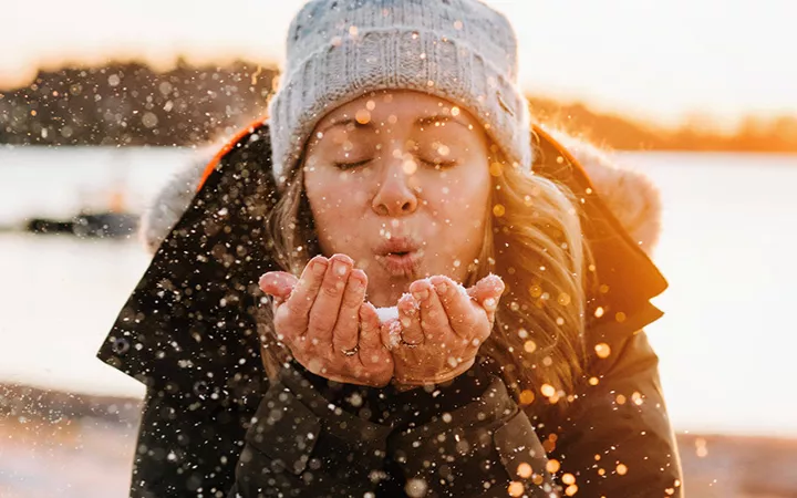 Eine Frau in Winterjacke & Mütze pustet Schnee in die Kamera 